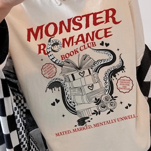 Op de afbeelding: Crèmekleurige sweatshirt met de tekst "MONSTER ROMANCE BOOK CLUB" in het rood. De afbeelding toont een open boek bovenop een stapel boeken, met tentakels, harten en de zinnen "ONE BED, SIX ARMS, NO REGRET, MEET NIGHT" en "COME FOR THE PLOT, STAY FOR THE TENTACLES".