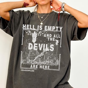 Op de afbeelding: Een donkergrijs oversized T-shirt met een witte grafische print die "De hel is leeg en alle duivels zijn hier -Shakespeare" zegt. De grafiek toont een skyline met een gevleugelde figuur in het midden.