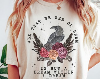 Könnte beinhalten: Ein cremefarbenes T-Shirt mit einem schwarzen Raben und einem floralen Design. Der Text lautet "All that we see or seem is but a dream within a dream" - Edgar Allan Poe.