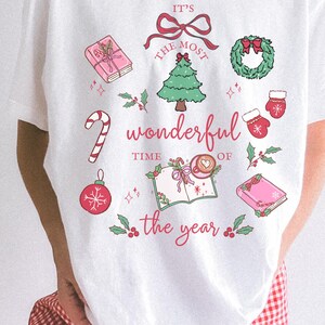 May include: White t-shirt with a festive Christmas design. The graphic includes a Christmas tree, wreath, mittens, and the text "It's the most wonderful time of the year" in red and pink. A candy cane and ornaments are also featured.