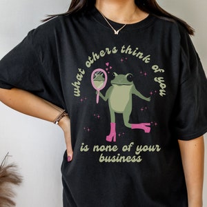 May include: Black oversized t-shirt with a green frog wearing pink boots and holding a mirror. The text on the shirt reads "What others think of you is none of your business."