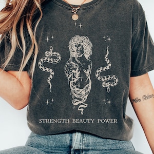 May include: A dark gray t-shirt with a graphic print of a woman with snakes and the text "STRENGTH BEAUTY POWER".