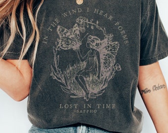 May include: A dark gray t-shirt with a graphic print of a fairy with wings, surrounded by flowers and leaves. The text "In the wind I hear poems Lost in time - Sappho" is printed on the shirt.