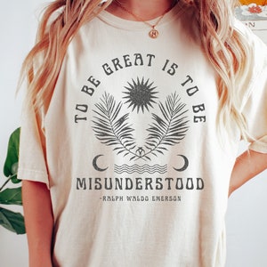 Op de afbeelding: Een cr&egrave;mekleurig T-shirt met een zwart grafisch ontwerp met de tekst "To be great is to be misunderstood - Ralph Waldo Emerson". De grafiek bevat een zon, palmbladeren en halve manen.