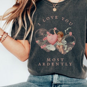 May include: A dark gray t-shirt with a graphic of a cherub holding a heart with an arrow through it. The text "I love you most ardently" is printed on the shirt.