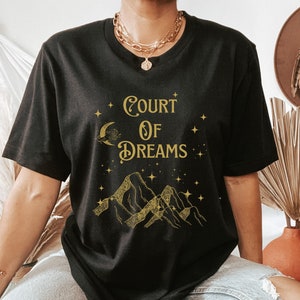 May include: A black t-shirt with a gold graphic design that says "Court of Dreams". The graphic features a crescent moon, stars, and mountains.
