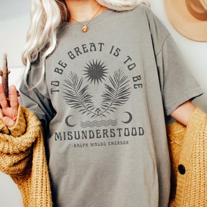 Op de afbeelding: Een grijs oversized t-shirt met een grafische print van een zon, maan en palmbladeren. De tekst op het shirt luidt "To be great is to be misunderstood - Ralph Waldo Emerson".