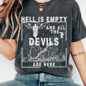 May include: A dark grey t-shirt with a white graphic print that reads "Hell is empty and all the devils are here". The graphic features a city skyline with a devil figure in the center.