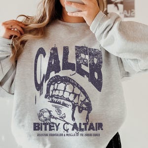 May include: A light gray sweatshirt with a dark blue graphic design. The design features the name "CALEB" above a stylized mouth with teeth, a rose, and the text "BITEY ALTAIR, CELESTIAL COUNCILLOR & MEMBER OF THE ZODIAC GUILD."