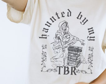 May include: A white t-shirt with a black graphic of a ghost holding a stack of books. The text "Haunted by my TBR" is printed on the shirt.