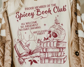 Puede incluir: Camiseta color crema con un gráfico burdeos que dice "Proud Member of the Spicey Book Club". El diseño incluye una calavera con vela, libros y rosas. La camiseta también dice "All Welcome, No Judgement, No Smut Shaming, Meets Every Friday."