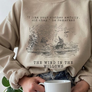 May include: A beige sweatshirt with a black and white illustration of characters from "The Wind in the Willows" book. The text "I like your clothes awfully. old chap, he remarked" and "THE WIND IN THE WILLOWS" is printed on the sweatshirt.