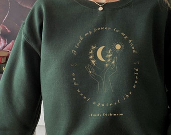 Può includere: Una felpa verde scuro con una stampa grafica dorata di mani che tengono una luna crescente e il sole. Il testo recita "Ho preso il mio potere nella mia mano e sono andato controcorrente - Emily Dickinson".