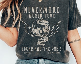 May include: A dark grey t-shirt with a graphic print of a skull with a snake wrapped around it. The text "NEVERMORE WORLD TOUR" is printed above the graphic and "EDGAR AND THE POE'S" is printed below the graphic. The text "22:66HR" and "9TH MAY 1982" are printed below the band name.