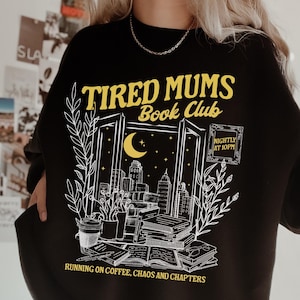 Puede incluir: Sudadera negra con un diseño gráfico blanco de una ventana con una ciudad, una luna creciente y libros. El texto "TIRED MUMS Book Club" está en amarillo con las palabras "NIGHTLY AT 10PM" en una fuente más pequeña. El texto "RUNNING ON COFFEE, CHAOS AND CHAPTERS" está en la parte inferior del diseño.