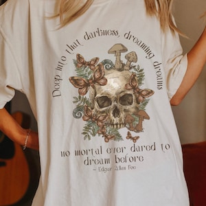 May include: A white t-shirt featuring a skull graphic surrounded by butterflies and mushrooms. The text "Deep into that darkness, dreaming dreams no mortal ever dared to dream before - Edgar Allan Poe" is printed on the shirt.