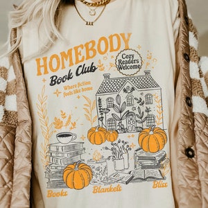 May include: A cream-colored t-shirt with a graphic design of a house with a sign that reads "Homebody Book Club" and "Cozy Readers Welcome". The graphic also includes pumpkins, books, and the words "Books", "Blankets", and "Bliss".