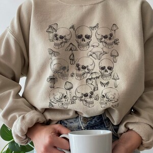 May include: A beige sweatshirt with a graphic print of skulls and mushrooms. The skulls are black and white and the mushrooms are brown. The sweatshirt has long sleeves and a crew neck.
