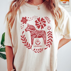 May include: A white t-shirt featuring a red Scandinavian Dala horse design. The horse is surrounded by floral patterns and leaves.