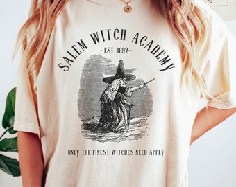 May include: A cream colored t-shirt with a black graphic of a witch with a pointed hat and a wand. The graphic is surrounded by the text "Salem Witch Academy - Est. 1692 -" and the text "Only the finest witches need apply".