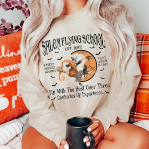 Op de afbeelding: Een beige sweatshirt met een grafische print van een heks die op een bezem voor een halve maan vliegt. De tekst luidt "Salem Flying School Est. 1892" en "Fly With The Best Over Three Centuries Of Experience".