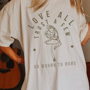 May include: White t-shirt with a brown frog sitting on a mushroom. The shirt has the text "Love All Trust A Few Do Wrong To None" printed on it.