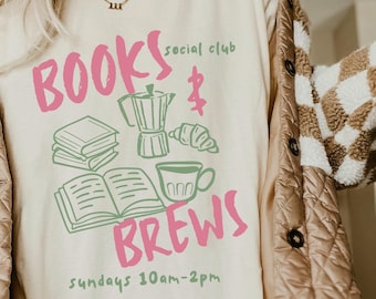 May include: A white t-shirt featuring a pink and green graphic design that reads "BOOKS & BREWS social club" and "Sundays 10am-2pm". The design includes illustrations of books, a coffee pot, a cup of coffee, and a croissant.