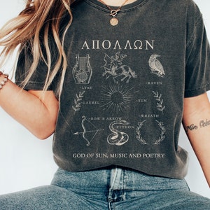 May include: A dark gray t-shirt with a graphic design featuring the Greek word "ΑΠΟΛΛΩΝ" and symbols representing the Greek god Apollo, including a lyre, laurel wreath, sun, raven, bow and arrow, and a python. The text "GOD OF SUN, MUSIC AND POETRY" is printed below the graphic.
