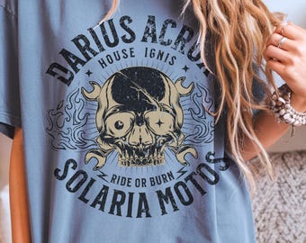 May include: A blue-grey t-shirt featuring a skull graphic with crossed wrenches and the text "DARIUS ACRO, HOUSE IGNIS, SOLARIA MOTOS, RIDE OR BURN". The design is distressed, giving it a vintage look. The shirt has short sleeves.