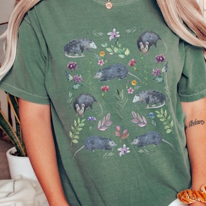 May include: Green t-shirt with a watercolor print of opossums and flowers. The opossums are in various poses, some are looking at the viewer, some are looking away. The flowers are in shades of pink, purple, and yellow.
