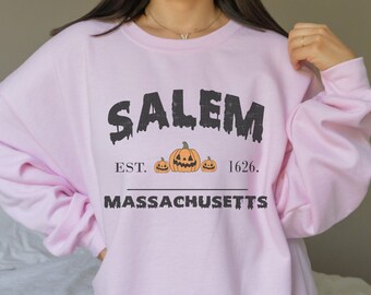 Op de afbeelding: Roze sweatshirt met een zwarte grafische print die "SALEM EST. 1626. MASSACHUSETTS" zegt met drie pompoengezichten.