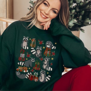 K&ouml;nnte beinhalten: Ein gr&uuml;ner Weihnachtspullover mit einem Muster aus Waschb&auml;ren, die Geschenke halten und Weihnachtsm&uuml;tzen tragen. Der Pullover hat einen Rundhalsausschnitt und lange &Auml;rmel.