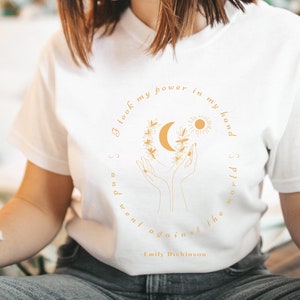 May include: White t-shirt with a gold graphic of hands holding a crescent moon and sun with a floral wreath. The text "I look my power in my hand and went against the world" is printed in gold with the author "Emily Dickinson" below.