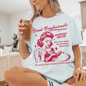 May include: A light blue t-shirt with a red graphic of a woman reading a book and crying. The text on the shirt reads "Book Boyfriends Anonymous Support Group Don't Cry Alone Meets Weekly Bring Wine Bonded by Books. United by Fictional Love."