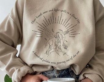 以下が含まれることがあります： 天使のグラフィックプリントが施されたベージュのトレーナー。テキストは「Their hands meet and light spills in a flood like a hundred golden urns pouring out of the sun」です。