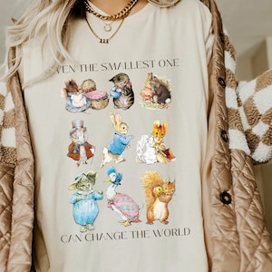 Könnte beinhalten: Ein beiges T-Shirt mit einem grafischen Druck von Figuren aus dem Kinderbuch "Die Geschichte vom Peter Hase". Der Text "Selbst der Kleinste kann die Welt verändern" ist unter den Figuren gedruckt.