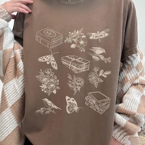 May include: A brown t-shirt with a white graphic print of various illustrations including books, flowers, butterflies, and dragonflies.