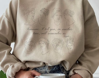 May include: A beige sweatshirt with a graphic print of several line drawings of human heads with a quote that reads "Someone, I tell you, in another time, will remember us - Sappho".