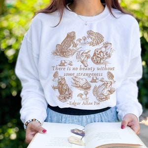 May include: White sweatshirt with a brown illustration of frogs and mushrooms. The text "There is no beauty without some strangeness - Edgar Allan Poe" is printed on the sweatshirt.