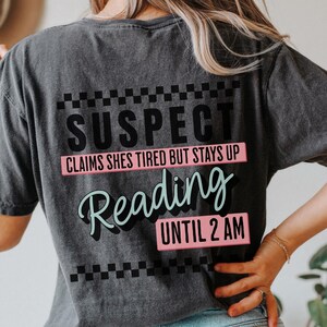May include: A black t-shirt with a white and black checkered pattern at the top and bottom. The text "SUSPECT" is in black, "CLAIMS SHES TIRED BUT STAYS UP" is in pink, "Reading" is in light blue, and "UNTIL 2 AM" is in pink.