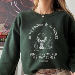 Op de afbeelding: Een donkergroen sweatshirt met een witte grafische print van een kristallen bol, kaarsen en de tekst "By the pricking of my thumbs, something wicked this way comes - Shakespeare".