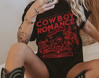 Peut inclure: T-shirt noir avec un motif rouge qui dit "Proud Member of the Cowboy Romance Book Club". Le motif représente un livre avec des fleurs et un cowboy à cheval.