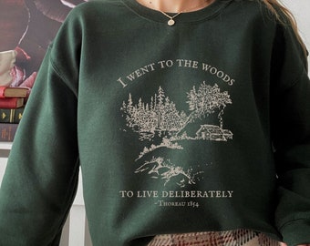 Pode incluir: Uma camisa de frio verde escura com uma estampa gráfica branca de uma cabana na floresta. O texto na camisa de frio diz "I went to the woods to live deliberately - Thoreau 1854".