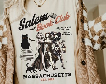 May include: A white t-shirt with a vintage-style graphic design featuring the text "Salem Book Club" with a black cat, three witches, and a skeleton. The text "All Witches Welcome Meets Monthly" and "Massachusetts Est. 1692" are also included in the design.