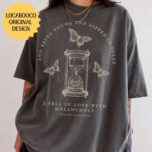 May include: A charcoal gray short-sleeve t-shirt featuring a graphic print of an hourglass with a skull inside, surrounded by three butterflies. The text reads: AND BEING YOUNG AND DIPPED IN FOLLY I FELL IN LOVE WITH MELANCHOLY - EDGAR ALLAN POE.
