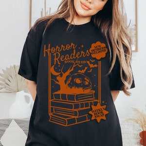 Puede incluir: Camiseta negra con un diseño gráfico naranja que presenta una calavera sobre una pila de libros. El texto "Horror Readers Book Club" y "Weekly Book Swap" están incluidos en el diseño.