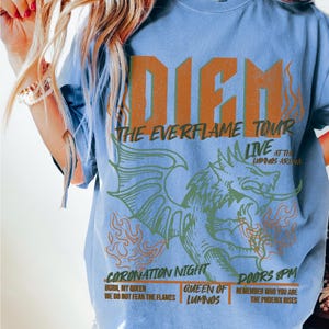 May include: A light blue t-shirt with the text "DIEM" in orange block letters. Below, it reads "THE EVERFLAME TOUR" and "LIVE AT THE LUMINOS ARENA". A dragon graphic is featured below the text. Additional text includes "CORONATION NIGHT" and "DOORS 8PM".