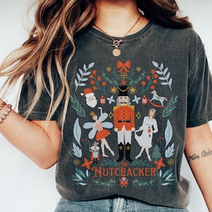 Puede incluir: Una camiseta gris oscuro con una ilustración colorida de El Cascanueces. La ilustración presenta un soldado cascanueces, una bailarina, un hada de azúcar y un caballo de balancín. El texto "The Nutcracker" está impreso debajo de la ilustración.
