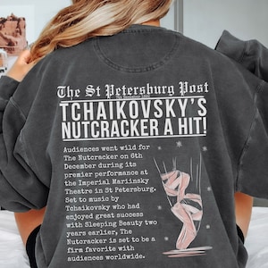 Puede incluir: Una sudadera gris con un diseño de periódico vintage que incluye el texto "The St Petersburg Post" y "Tchaikovsky's Nutcracker a Hit!" El texto describe el éxito de la representación de ballet. Una ilustración de bailarina rosa se encuentra en el lado derecho de la sudadera.