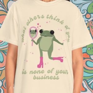 May include: A light beige t-shirt with a green frog wearing pink boots and holding a pink mirror. The frog is looking at its reflection in the mirror. The text on the shirt reads "what others think of you is none of your business".